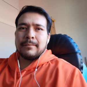 Retrato de una persona con sudadera naranja, sentada en una silla negra, en un entorno interior bien iluminado. La persona usa auriculares y sonríe suavemente.